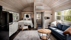 A cabin interior with a bed, sofa, TV, and kitchenette. - Modern Tiny Cabin with Game Room & Fire Pit in West Virginia (Great Cacapon)