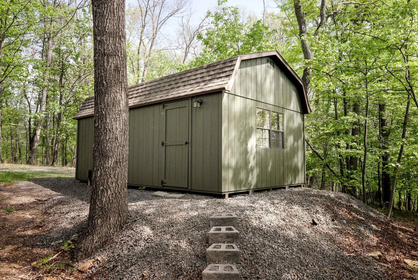 A wooden shed with a sloped roof, a door, and a window, surrounded by trees and gravel.