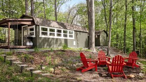 A cabin with a deck, a covered outdoor area, and a small shed in a wooded setting. - Modern Tiny Cabin with Game Room & Fire Pit in West Virginia (Great Cacapon)