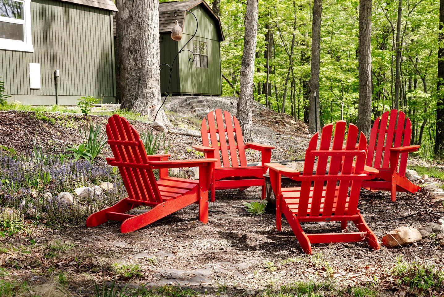 A wooden deck with three red Adirondack chairs, a small table, and a green shed in a forested area.