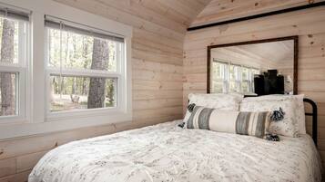 A bedroom with a bed, two windows, a mirror, and a view of trees.