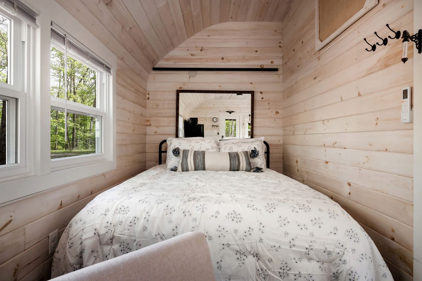 A wooden cabin bedroom with a bed, two windows, a mirror, and a wall-mounted light fixture.