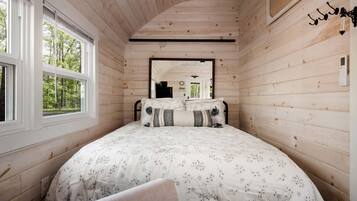 A wooden cabin bedroom with a bed, two windows, a mirror, and a wall-mounted light fixture.