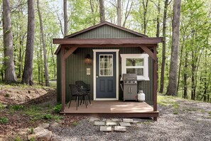 Eine kleine Holzhütte mit Tür, Fenster, Grill und zwei Stühlen davor.