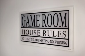 Ein Schild an einer weißen Wand mit der Aufschrift 'GAME ROOM HOUSE RULES' und 'KEIN BETRUG - KEINE SCHLAGEKÄMPFE - KEINE NÖRGELEI'.