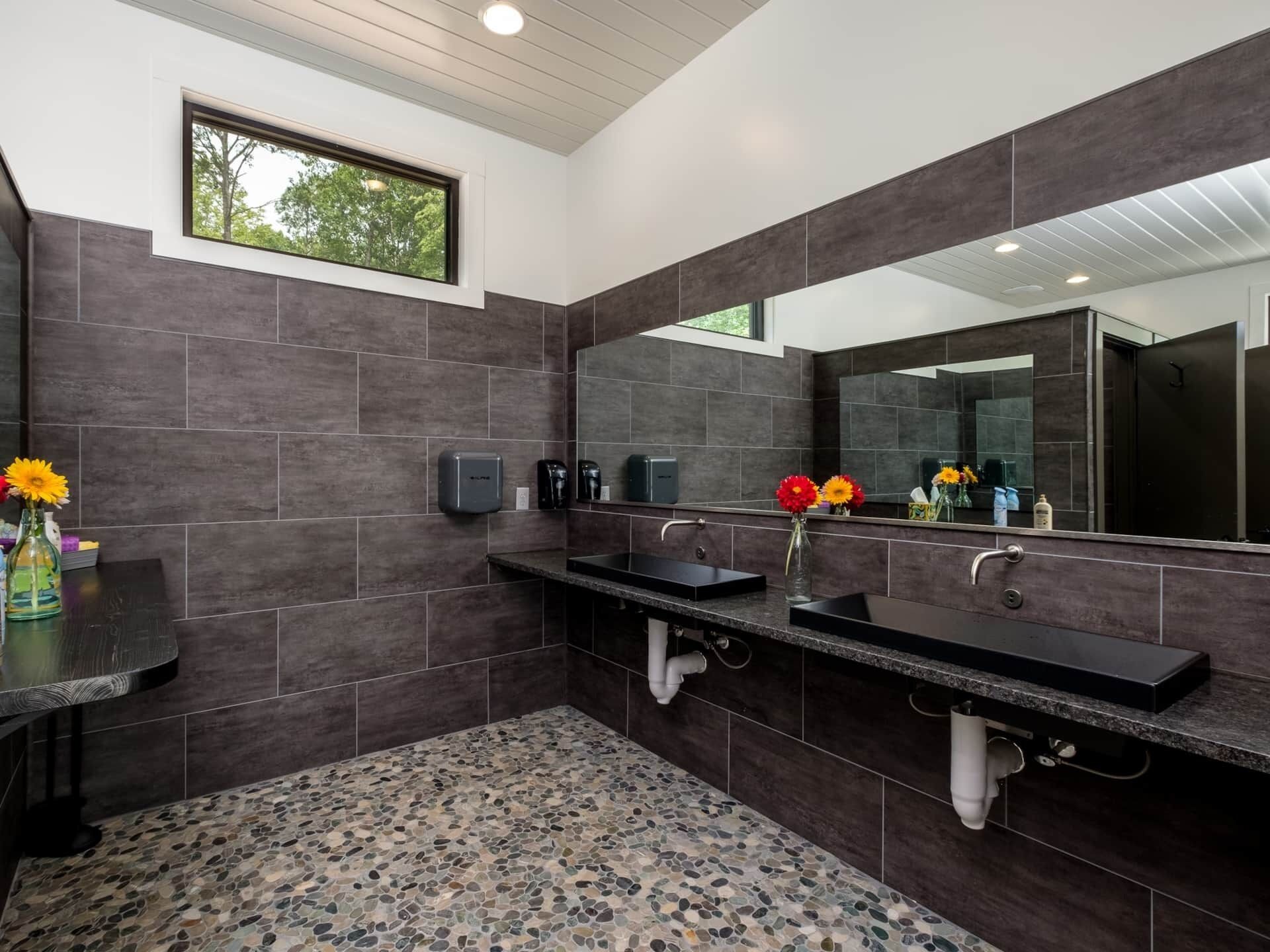 Une salle de bain moderne avec des carreaux de couleur foncée, deux lavabos, un grand miroir et une fenêtre.