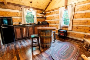 A log cabin interior with a kitchen area featuring a black refrigerator, wooden cabinets, a round wooden table, and a small stove.