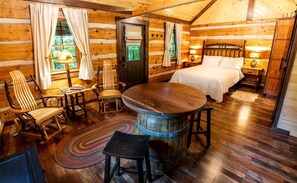 A log cabin room with a bed, a wooden table, a chair, a small rug, and a wooden barrel.