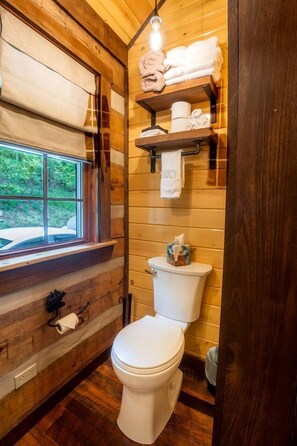 Ein Badezimmer einer Holzhütte mit Toilette, Regalen mit Handtüchern, einem Fenster und einer Deckenleuchte.
