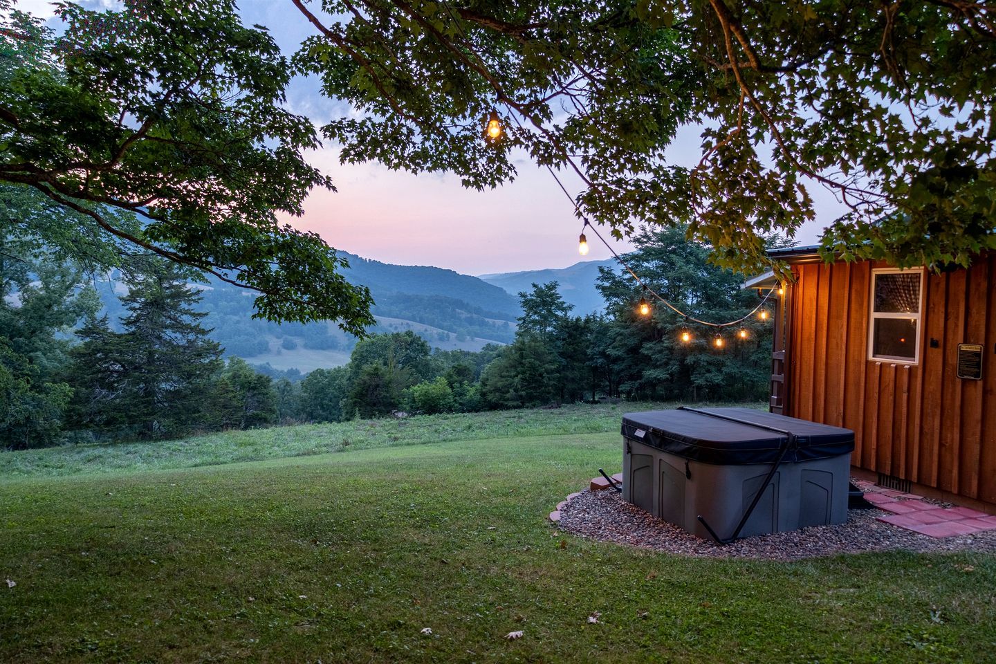 A hot tub in a grassy area with a wooden cabin and string lights.