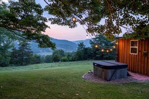 A hot tub in a grassy area with a wooden cabin and string lights.