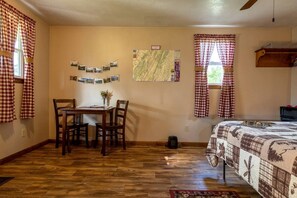 A room with a wooden floor, a bed with a patterned bedspread, a dining table with chairs, a map on the wall, and checkered curtains.