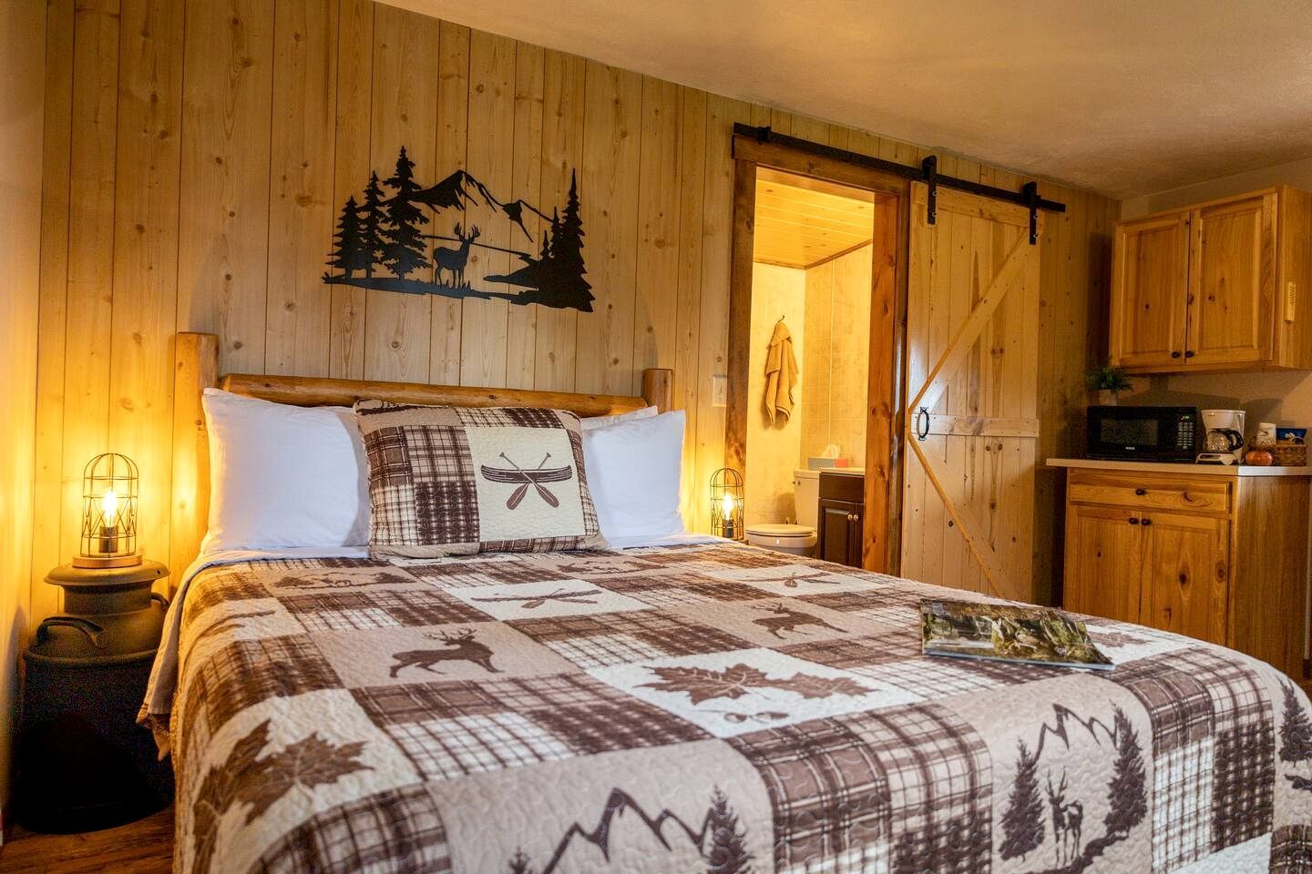 A wooden room with a bed, a wooden wall with a mountain and deer silhouette, a kitchen area, and a sliding door.