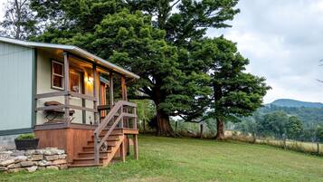 A cabin with a wooden deck, a staircase, and a large tree in the background.