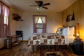 A bedroom with a bed, a ceiling fan, a window with checkered curtains, a bedside table, and a lamp.