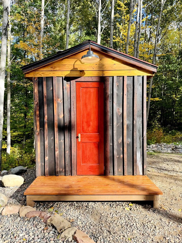 Een houten tuinhuisje met een rode deur en een lamp erboven, gelegen in een bebost gebied met een grindpad.
