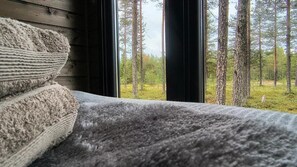 A bed with a grey blanket and a view of a forest through a window.