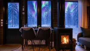 A cozy room with a wood-burning stove, two chairs, a table, and a view of the aurora borealis through large windows. - Borealis (Muonio)