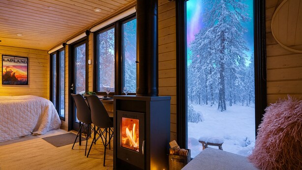 A cozy cabin interior with a wood-burning stove, large windows, and a bed. - Borealis (Muonio)