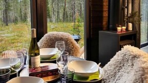 A dining table set with plates, glasses, and a bottle of wine, with a view of a forest through large windows. - Borealis (Muonio)