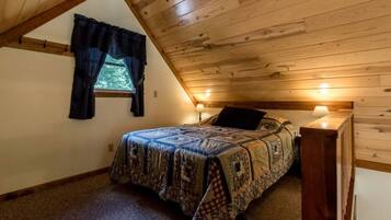 Een slaapkamer met een schuine kap, een bed met een geborduurde spreideken, twee bedlampjes en een raam met gordijnen.