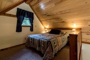 Ein Schlafzimmer mit schrägen Wänden, einem Bett mit gemustertem Überwurf, zwei Nachttischlampen und einem Fenster mit Vorhängen.