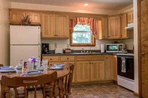 A kitchen with wooden cabinets, a refrigerator, a microwave, a stove, and a dining table set with plates and glasses. - Secluded West Virginia Cabin Rental on the Edge of Monongahela National Forest (Riverton)