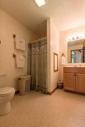 A bathroom with a toilet, a shower curtain, a sink with a mirror, and a trash bin. - Secluded West Virginia Cabin Rental on the Edge of Monongahela National Forest (Riverton)