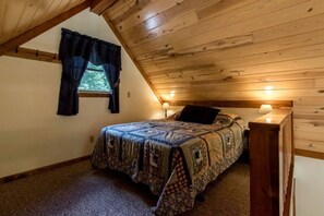 A bedroom with a sloped ceiling, a bed with a patterned bedspread, two bedside lamps, and a window with curtains.