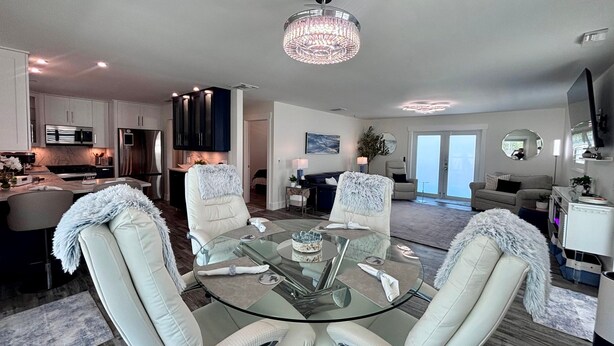 A modern living room with a glass dining table, white leather chairs, a chandelier, and a kitchen in the background. - Tropical Hideaway in Fort Myers Beach (Fort Myers Beach)