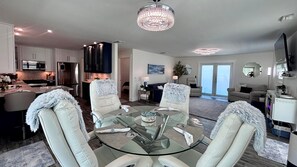 A modern living room with a glass dining table, white leather chairs, a chandelier, and a kitchen in the background. - Tropical Hideaway in Fort Myers Beach (Fort Myers Beach)