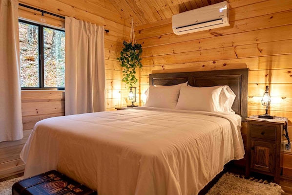 Un dormitorio de cabaña de madera con cama, cortinas, aire acondicionado, una planta colgante y una mesita de noche con lámpara.