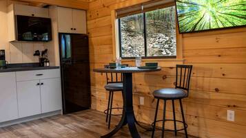 A wooden cabin interior with a kitchen, a dining table with two chairs, and a large window with a forest view.