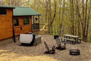 A cabin with a covered outdoor seating area, a fire pit, and a view of the surrounding forest.