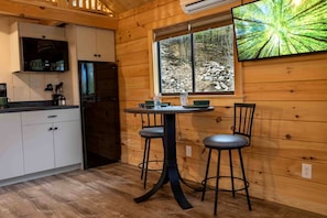 Interior de una cabaña de madera con cocina, una mesa de comedor con dos sillas y un ventanal con vistas a un bosque.