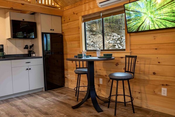 Ein Holzhütten-Interieur mit einer Küche, einem Esstisch mit zwei Stühlen und einem großen Fenster mit Blick auf einen Wald.