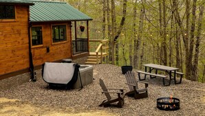 A cabin with a covered outdoor seating area, a fire pit, and a view of the surrounding forest.