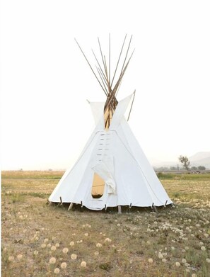 Una tienda tipi blanca con entrada central, ubicada en un campo herboso con vegetación escasa.