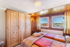 A bedroom with a wooden wardrobe, a bed with patterned bedding, a window with curtains, and a view of the outside.