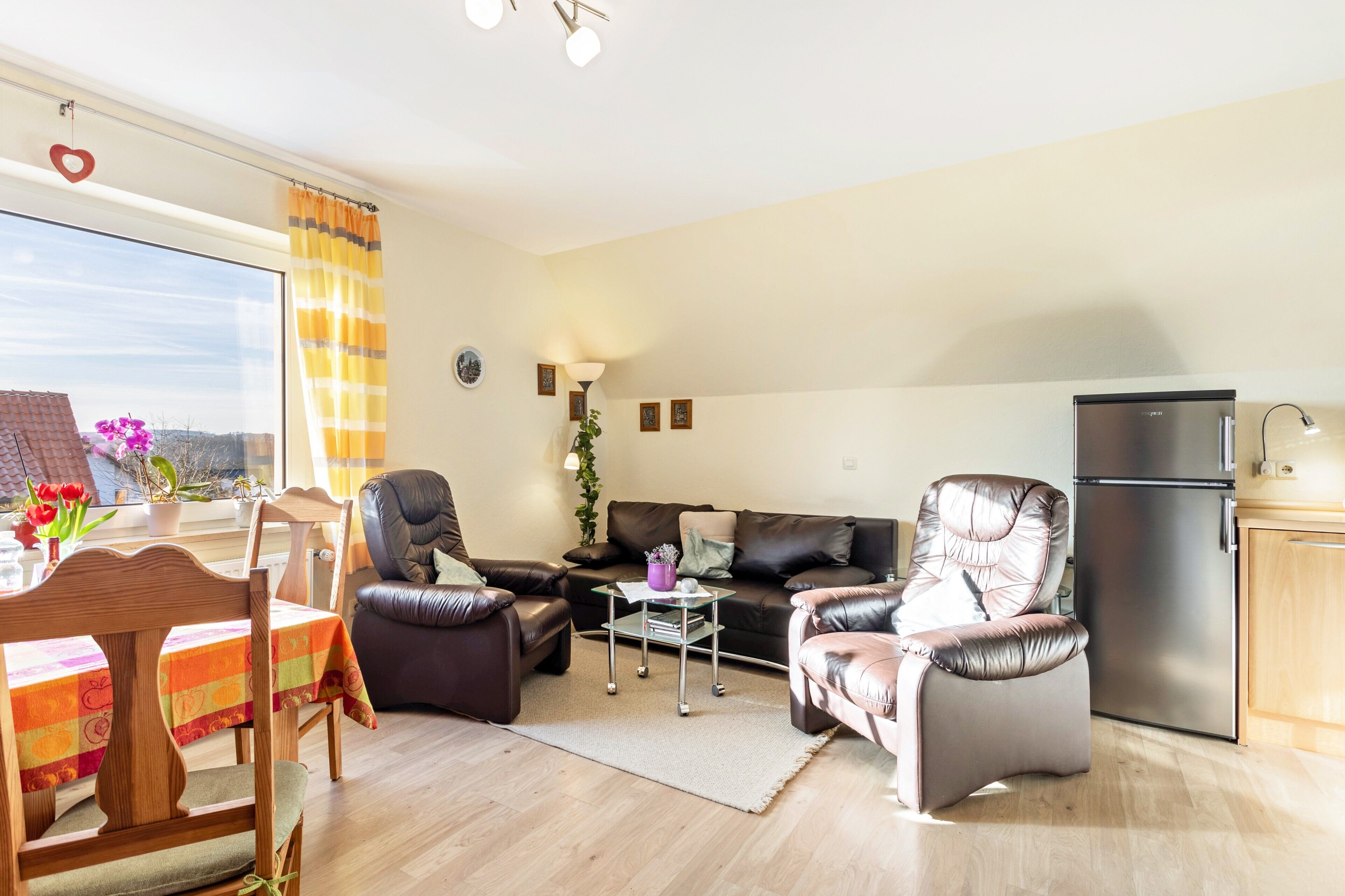 A modern living room with a dining table, two leather sofas, a glass coffee table, a refrigerator, and a kitchen area.