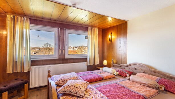 Une chambre avec un plafond en bois, un lit avec une literie à motifs, un banc, une fenêtre avec des rideaux et une lampe fixée au mur.
