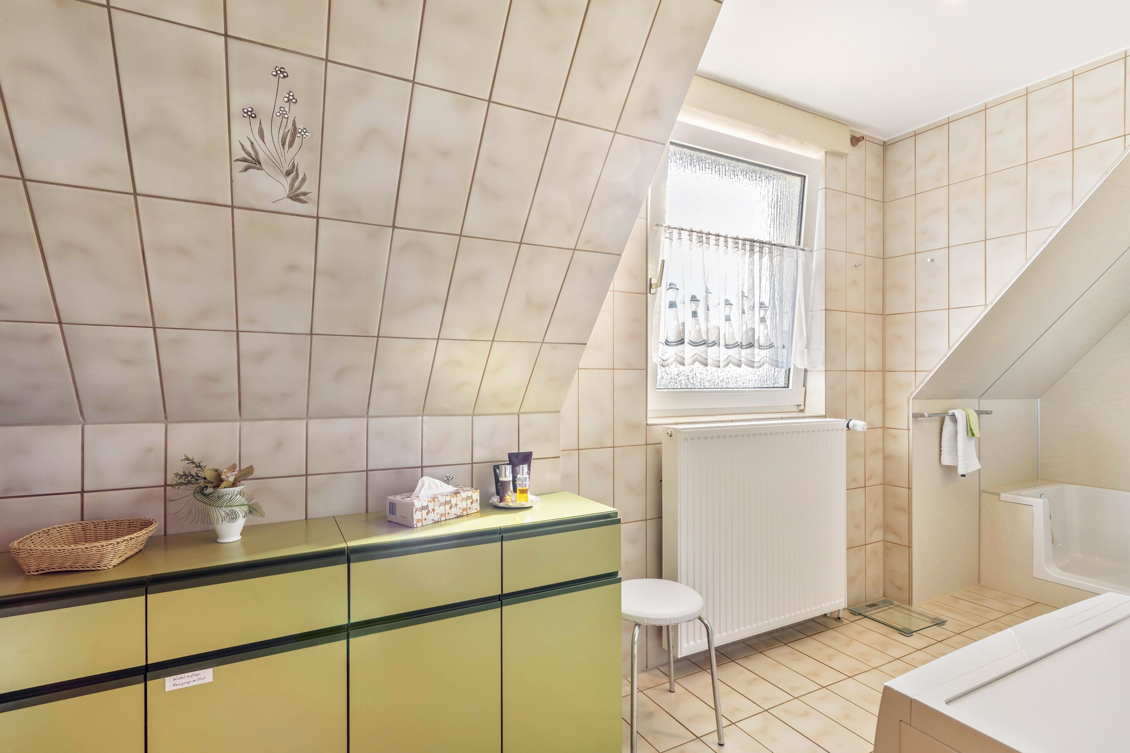 Et badeværelse med badekar, et vindue med gardiner, en radiator og en grøn kommode med en plante.