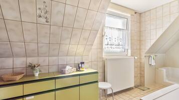 Et badeværelse med badekar, et vindue med gardiner, en radiator og en grøn kommode med en plante.