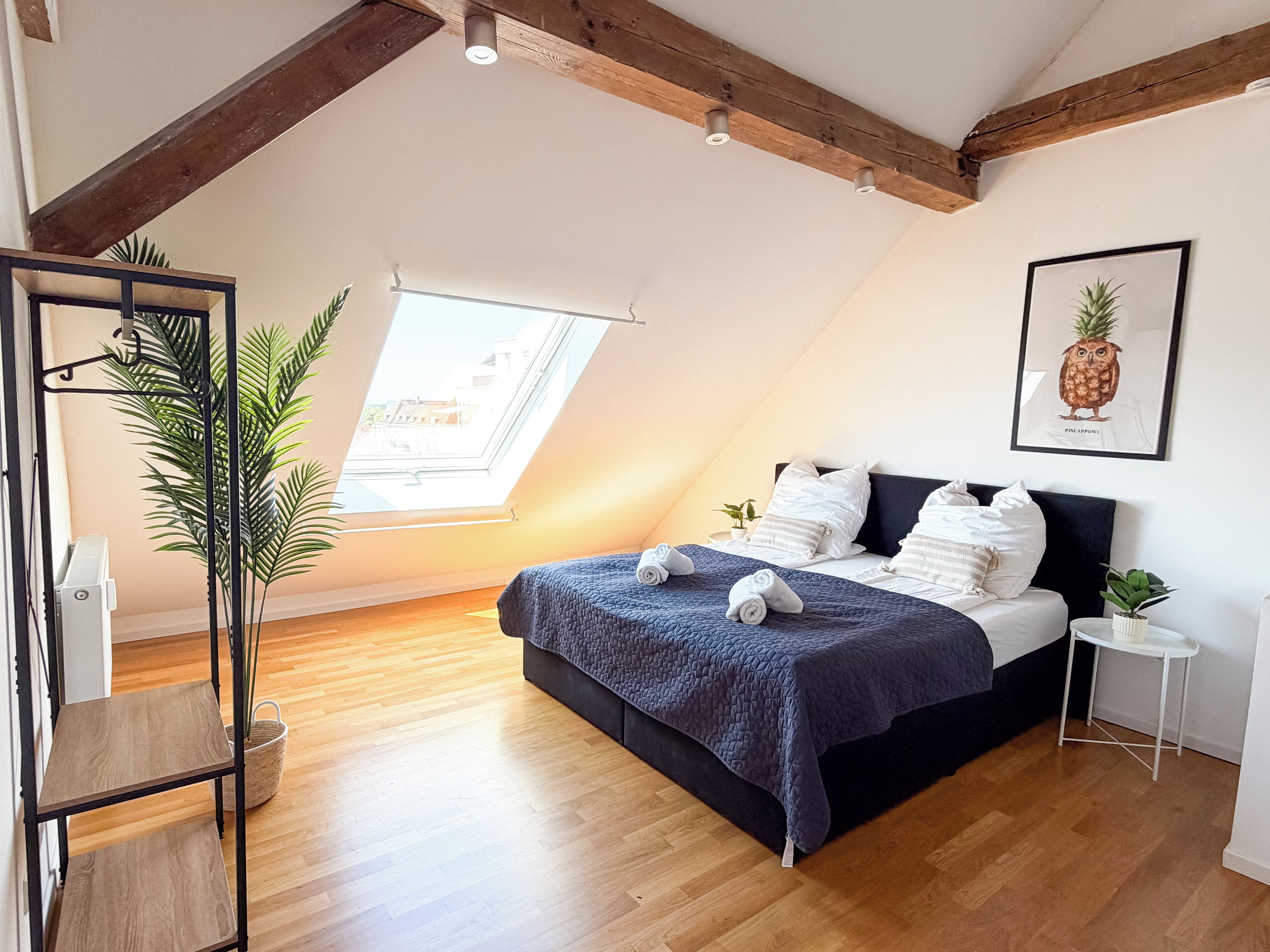 A bedroom with a skylight, a bed with a dark headboard, a framed pineapple artwork, and a wooden floor.