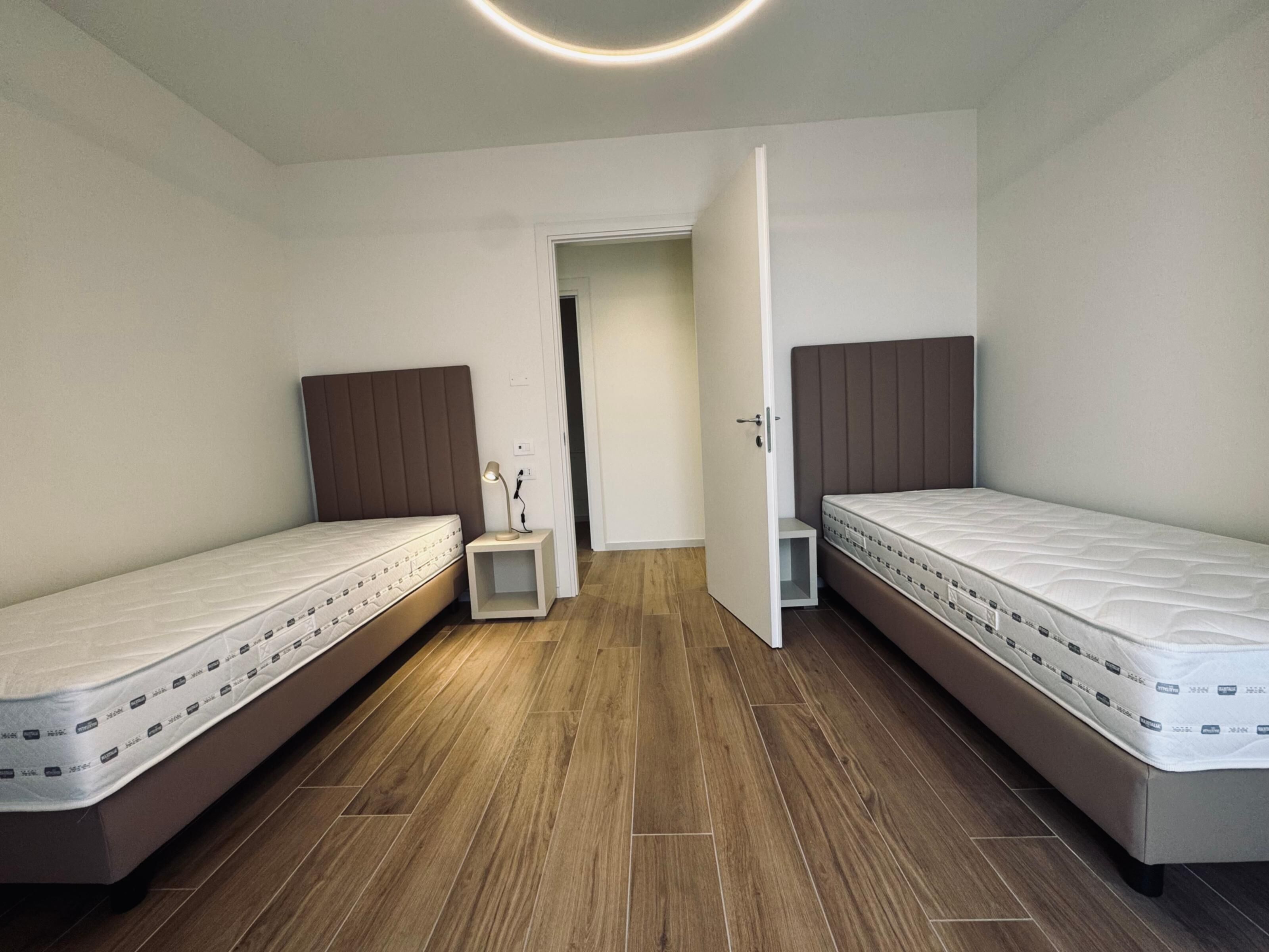 Un dormitorio con dos camas individuales, una mesita de noche con lámpara y una puerta que da a otra habitación.