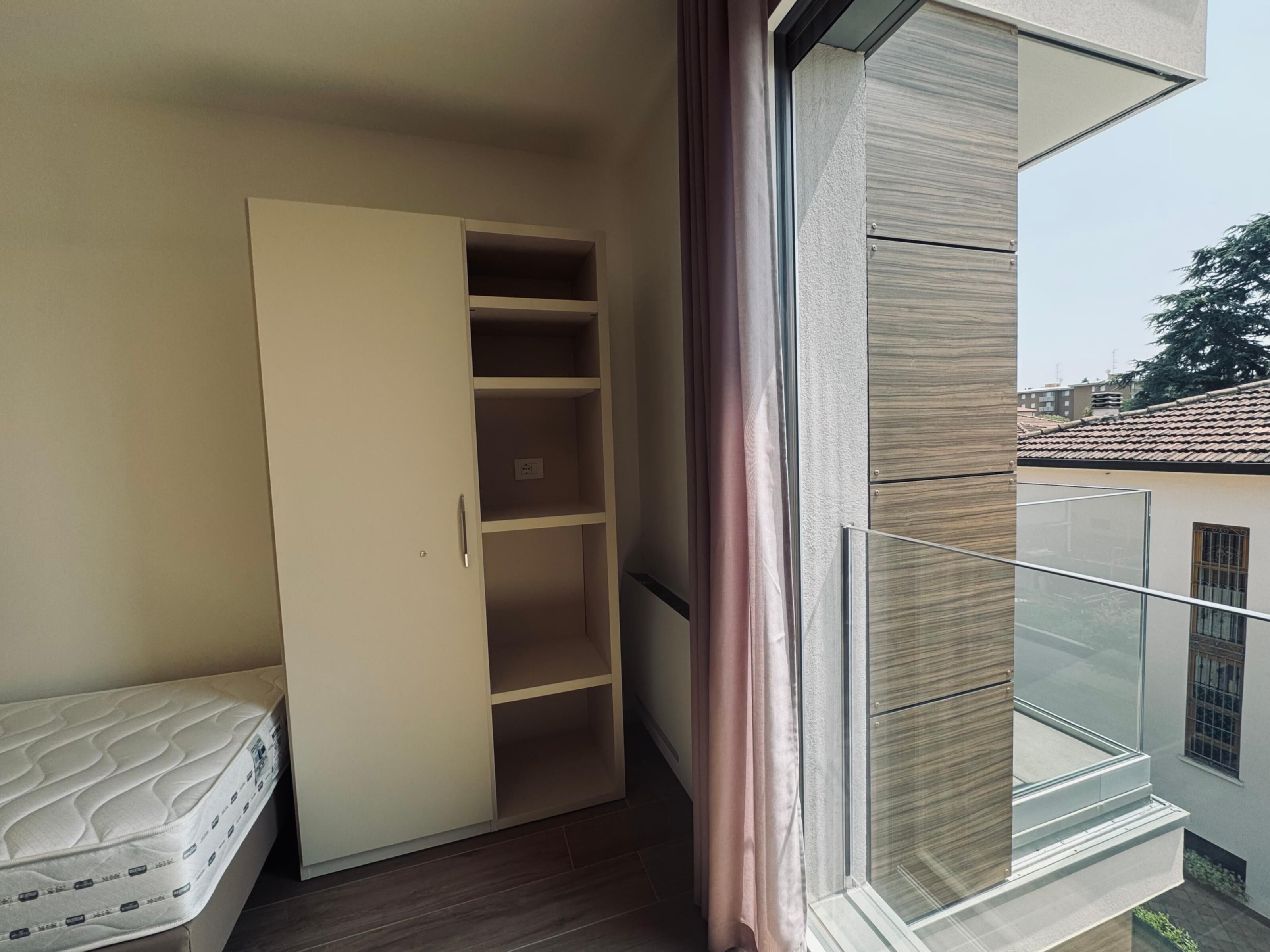 Une petite pièce comprenant un lit, une armoire et une fenêtre donnant sur des immeubles et des arbres.