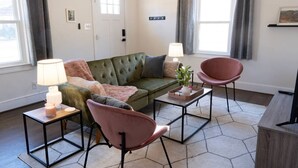 Un salon moderne avec un canapé vert, deux fauteuils, une table basse et un téléviseur. On y trouve deux lampes, une plante et une petite étagère avec un tableau.