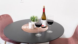Une table ronde à la surface noire, deux chaises roses, une bouteille de vin, deux verres à vin, une petite plante, et des salières et poivrières.