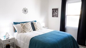 Une chambre à coucher bien rangée, avec un lit, une table de chevet, une lampe et une fenêtre avec des rideaux.