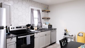 Une cuisine moderne avec un four noir, des armoires blanches et un lave-vaisselle gris. On y trouve une fenêtre avec des rideaux gris, un four à micro-ondes posé sur le plan de travail et une poubelle jaune.